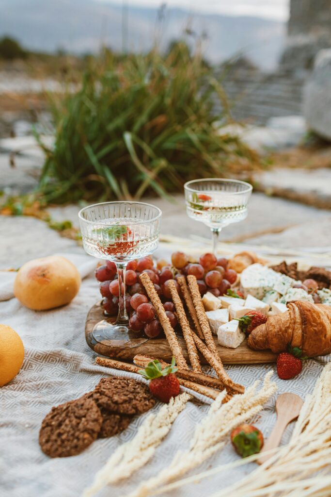 pexels-photo-6594962-6594962 Charming picnic spread with wine glasses, cheese, grapes, and snacks in a rustic outdoor setting.
